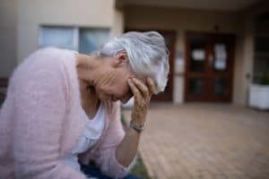 Side view of depressed senior woman sitting on bench with head in hand against nursing home.