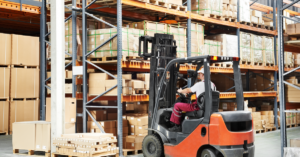 Warehouse worker driver in uniform loading cardboard boxes by forklift stacker loader.